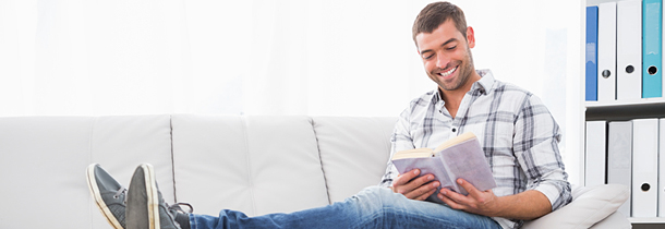 Junger sportlicher Mann sitzt auf dem Sofa, liest ein Buch und geniesst die Entspannung