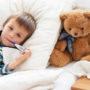 Kleiner Junge liegt im Bett mit Thermometer im Mund und Teddybär an seiner Seite