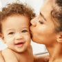 Eine junge Mutter küsst ihr Baby nach dem Stillen währenddem das Baby lächelnd zum Betrachter blickt
